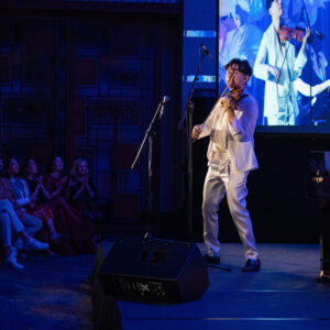 Violinist in white performing solo on a blue-lit stage, a close-up projected behind as guests watch nearby