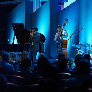 Jazz quartet in blue light with harmonica, double bass, piano and drums playing to a seated evening audience