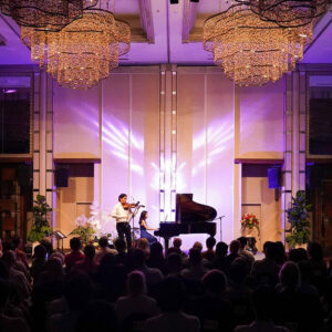 Violinist and young pianist performing a duet on a bright stage beneath sparkling chandeliers before a packed hall