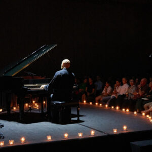 Pianist at a grand piano on a candle-lined stage, warm flames glowing before a seated audience in darkness