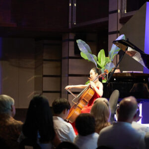 Cellist in a red gown playing beside a grand piano, framed by tropical foliage before a close-seated audience