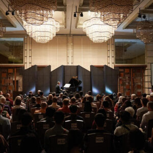 Pianist at a grand piano on a screened stage beneath glittering chandeliers, a full audience watching