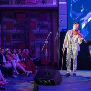 Violinist in white taking a bow on a violet-lit stage, a piano close-up projected behind and guests seated beside him
