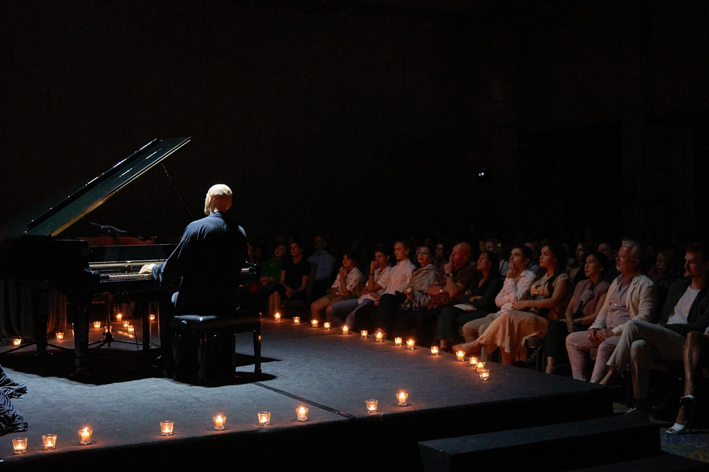 Pianist at a grand piano on a dark stage lit by rows of candles, audience seated close by